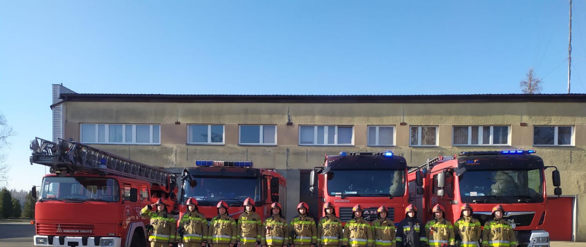 Zdjęcie przedstawia trzynastu strażaków w ubraniach specjalnych stojących w szeregu przed ustawionymi obok budynku Jednostki Ratowniczo - Gaśniczej Państwowej Straży Pożarnej w Wieruszowie samochodami pożarniczymi.