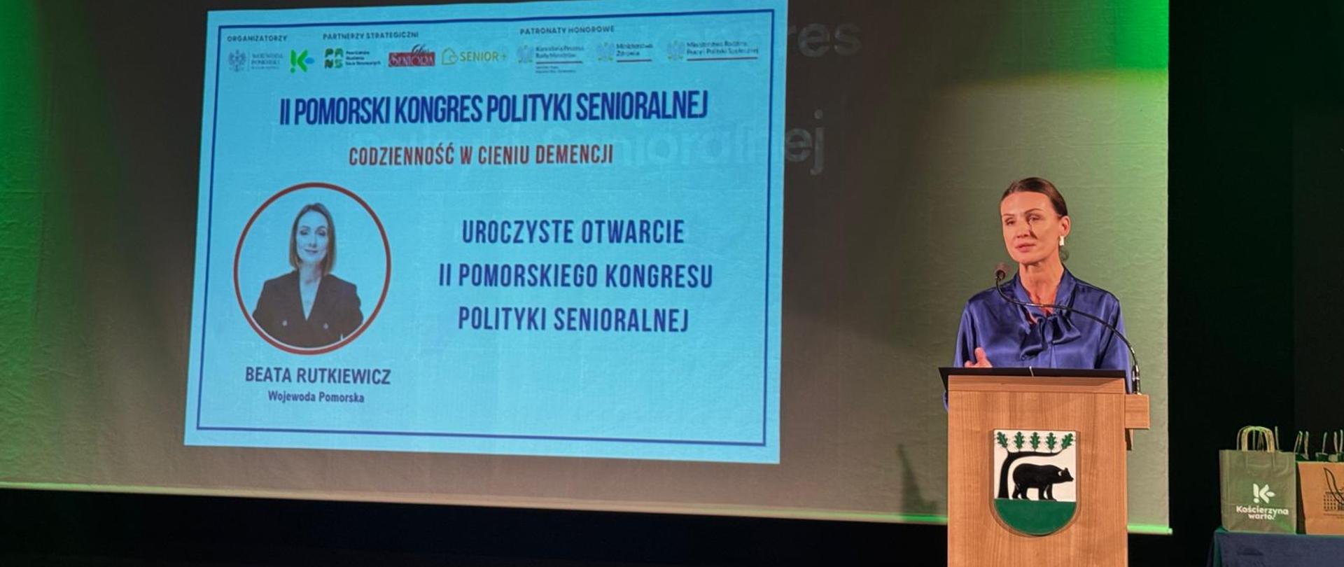 kobieta stoi za mównicą, na scenie, w tle tablica z napisem pomorski kongres senioralny
