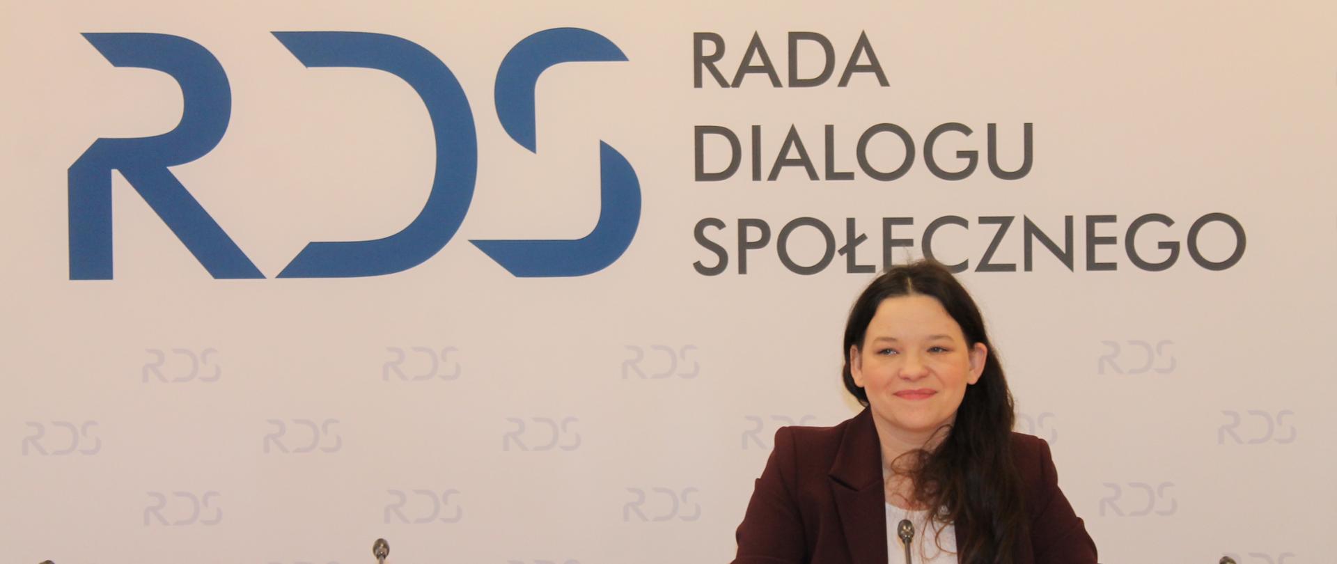 Na zdjęciu widnieje stół prezydialny za którym siedzi Pani Katarzyna Nowakowska, Podsekretarz Stanu w Ministerstwie Rodziny, Pracy i Polityki Społecznej. W tle znajduje się baner z napisem Rada Dialogu Społecznego