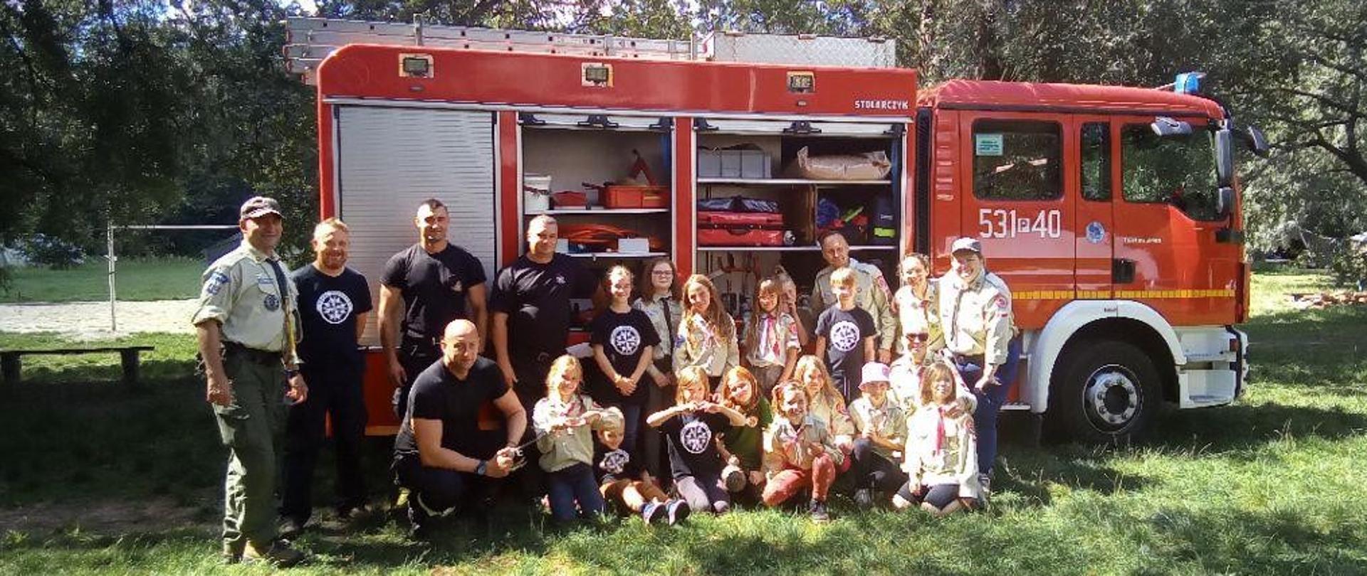 Harcerze oraz funkcjonariusze PSP na obozie harcerskim, w tle samochód strażacki