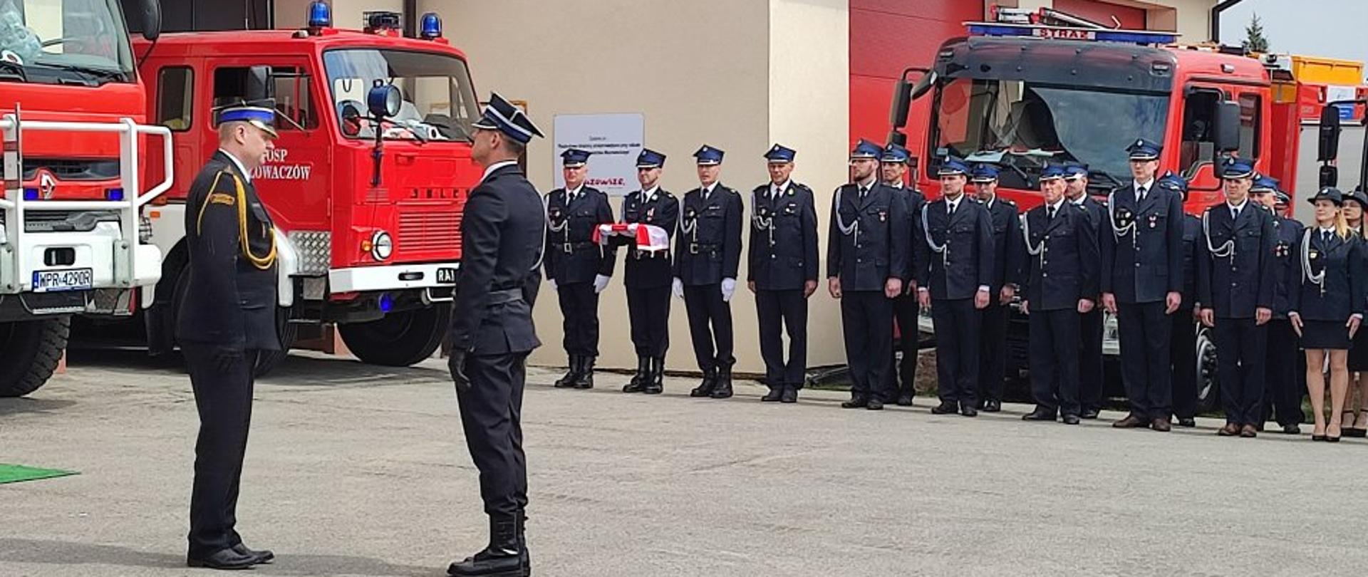 na pierwszym planie Komendant Powiatowy PSP w Kozienicach bryg. Kamil Bieńkowski odbiera meldunek od dowódcy uroczystości druha Wiktora Czubaja, w tle druhowie OSP stojący na placu przy samochodach strażackich