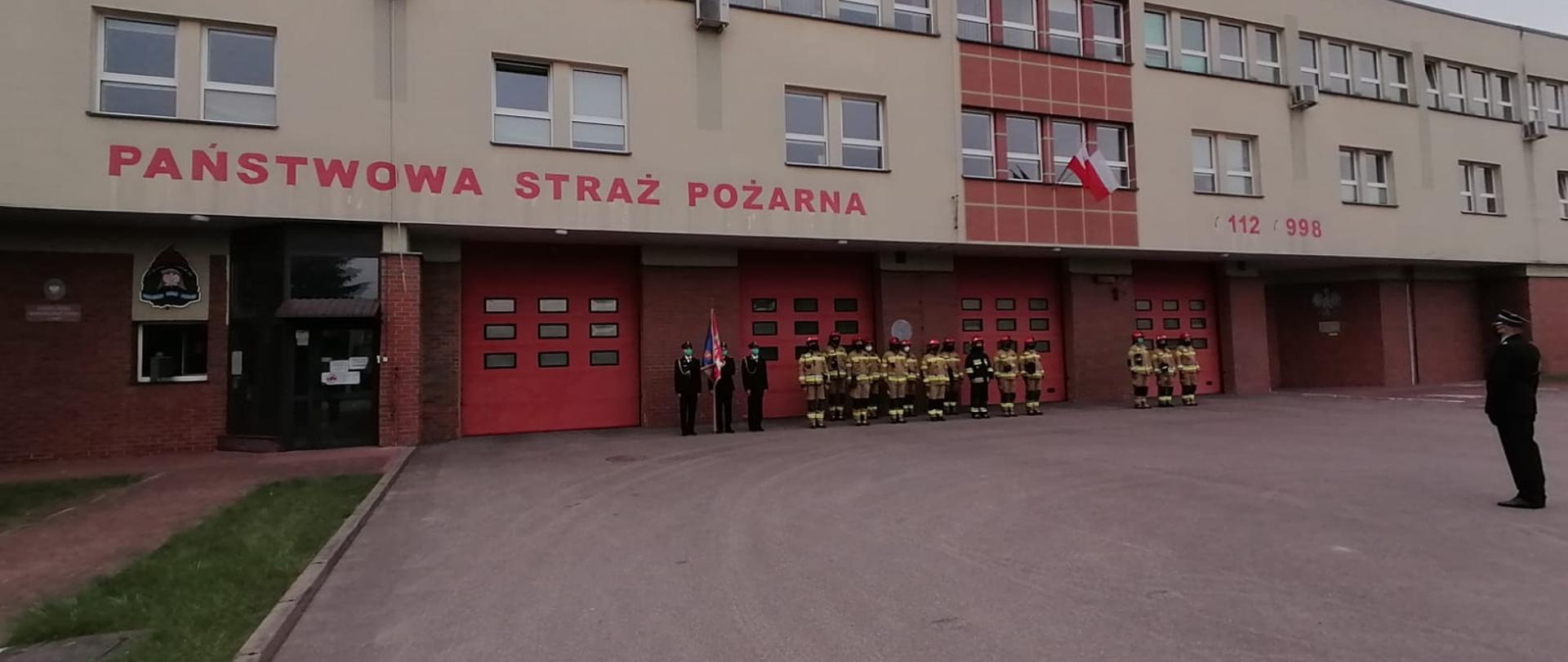 Zbiórka strażaków Jednostki Ratowniczo - Gaśniczej w Łomży w obecności dowódcy JRG podczas uroczystego apelu z okazji Dnia Flagi Rzeczypospolitej Polskiej. Ustawieni strażacy w pododdziałach przed głównym wejściem komendy.
