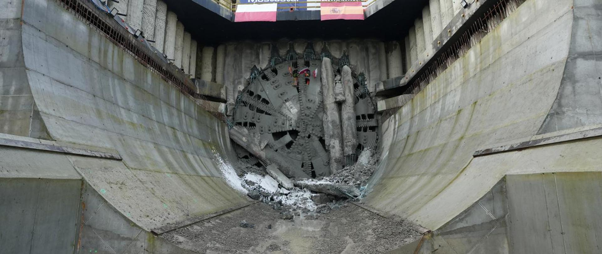Widok tarczy TBM (Tunnel Boring Machine) przebijającej się przez betonową ścianę w komorze startowej lub odbiorczej tunelu. Wokół tarczy widoczne są fragmenty rozkruszonego betonu i ziemi, a ściany komory są wykonane z żelbetu. Na górze widać fragment konstrukcji z banerami.