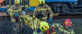 Na zdjęciu znajdują się strażacy w ubraniach specjalnych ewakuujący po nasypie kolejowym osobę poszkodowaną, w tle wagon kolejowy pasażerski.