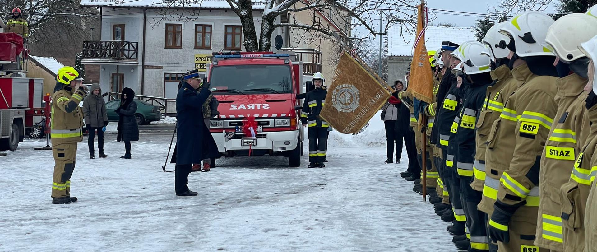 W centralnej części znajduje się lekki pojazd ratowniczo gaśniczy. Po prawej stronie w szeregu stoją w umundurowani strażacy, na przeciwko nich stoi Zastępca Warmińsko Mazurskiego Komendanta Wojewódzkiego. W tle zabudowania miejskie.