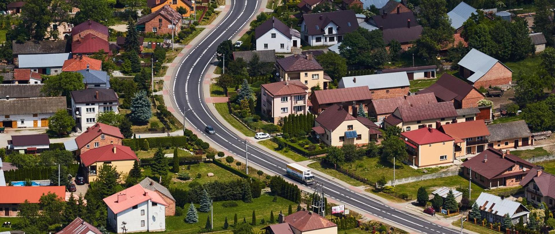 Widok z lotu ptaka na niewielką miejscowość. Przez środek zdjęcia biegnie szeroka, nowa droga o gładkiej, ciemnej nawierzchni, łagodnie wijąca się między zabudowaniami. Po obu stronach znajdują się jednorodzinne domy z dachami w różnych kolorach – czerwonych, brązowych i szarych – otoczone ogrodami, drzewami i terenami zielonymi. Na drodze widocznych jest kilka pojazdów, w tym samochody osobowe i ciężarówka. Całość sprawia wrażenie zadbanej, spokojnej podmiejskiej okolicy z dużą ilością zieleni.