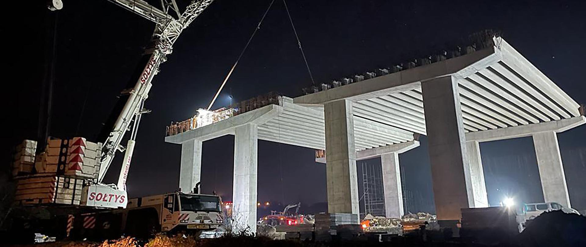 zdjęcie nocne, fragment wysokiego wiaduktu w budowie, obok stoi dźwig