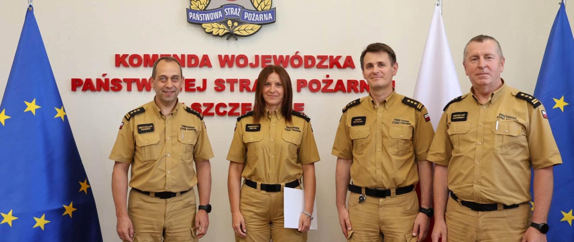 Na zdjęciu pięć osób – czterech funkcjonariuszy i jedna funkcjonariuszka Państwowej Straży Pożarnej – stoi w jednym rzędzie podczas uroczystości w siedzibie komendy. Wszyscy ubrani są w służbowe mundury w kolorze piaskowym. Osoba stojąca pośrodku trzyma białą teczkę z dokumentem.