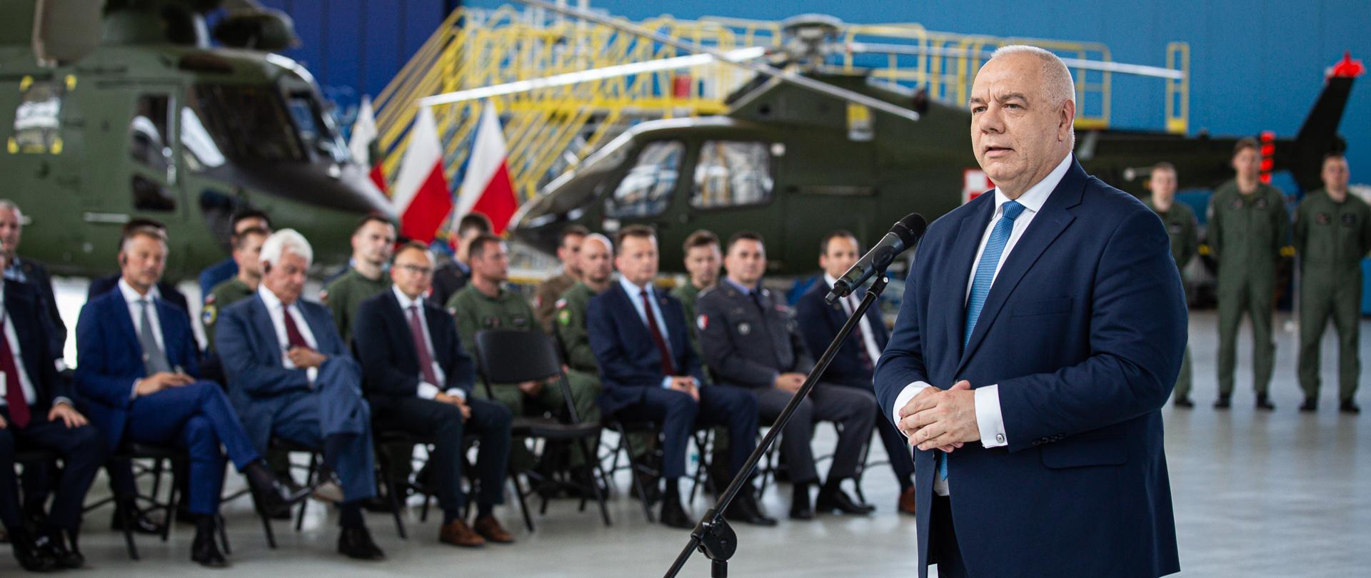 Szerokie ujęcie. Wicepremier Jacek Sasin stoi za mikrofonem. W tle śmigłowce i wojsko. Po lewej stronie siedzą uczestnicy spotkania.