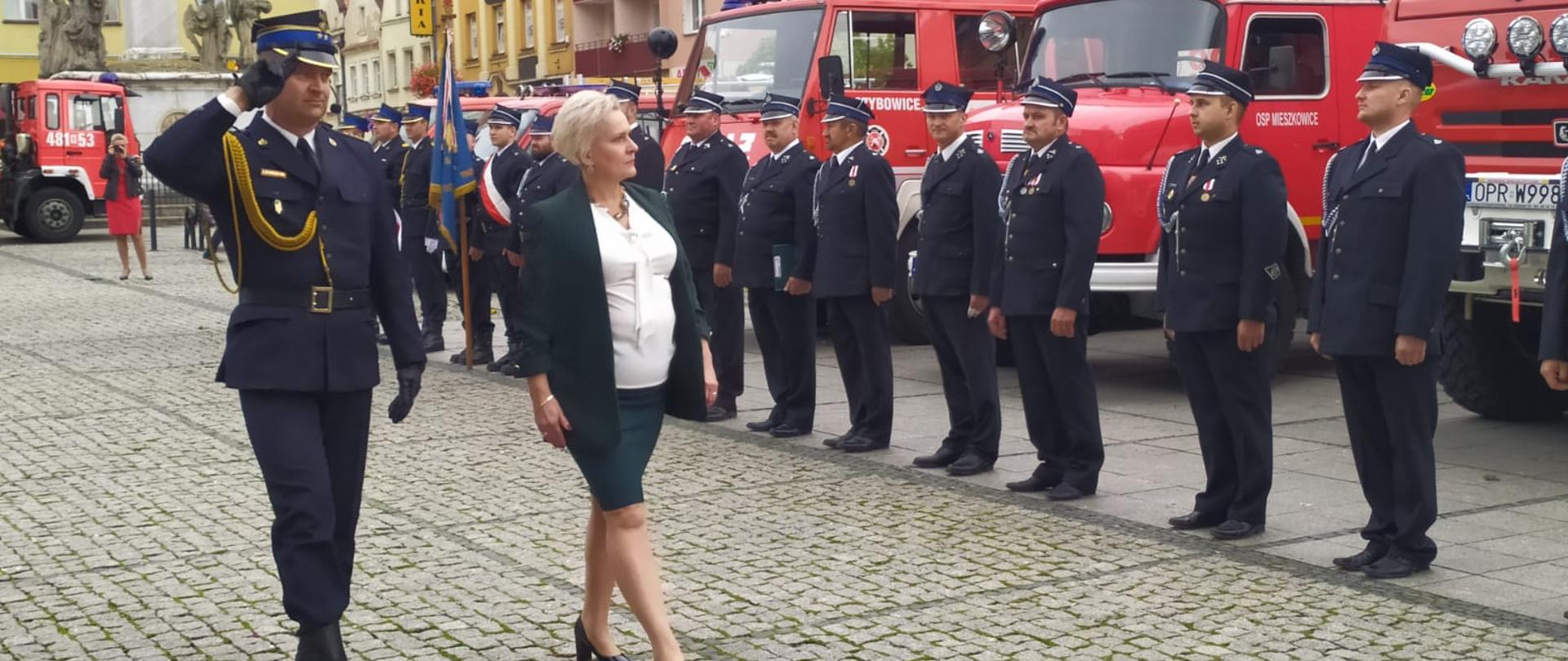 Teresa Barańska podczas przeglądu pododdziałów gotowych do uroczystej zbiórki. W tle ustawieni strażacy, za nimi wozy ratowniczo-gaśnicze. 