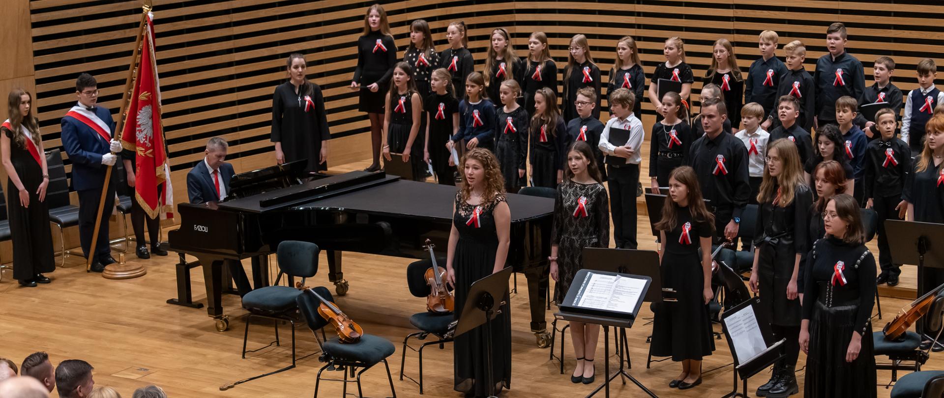 Na estradzie w dwóch rzędach stoi ok. trzydzieścioro dzieci, przed nimi stoi osiem osób - wszyscy śpiewają. Po lewej stronie mężczyzna gra na fortepianie, jeszcze bardziej na lewo przy ścianie stoi dziewczyna i chłopak, który trzyma sztandar. Z przodu id tyłu widać stojącą publiczność.