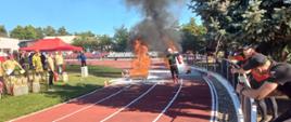 Zawodnik biegnie po torze. Przed zawodnikiem ogień. Zawodnik trzyma czerwoną gaśnic1)ę. Zdjęcie zrobione w ciągu dnia. Niebo błękitne.