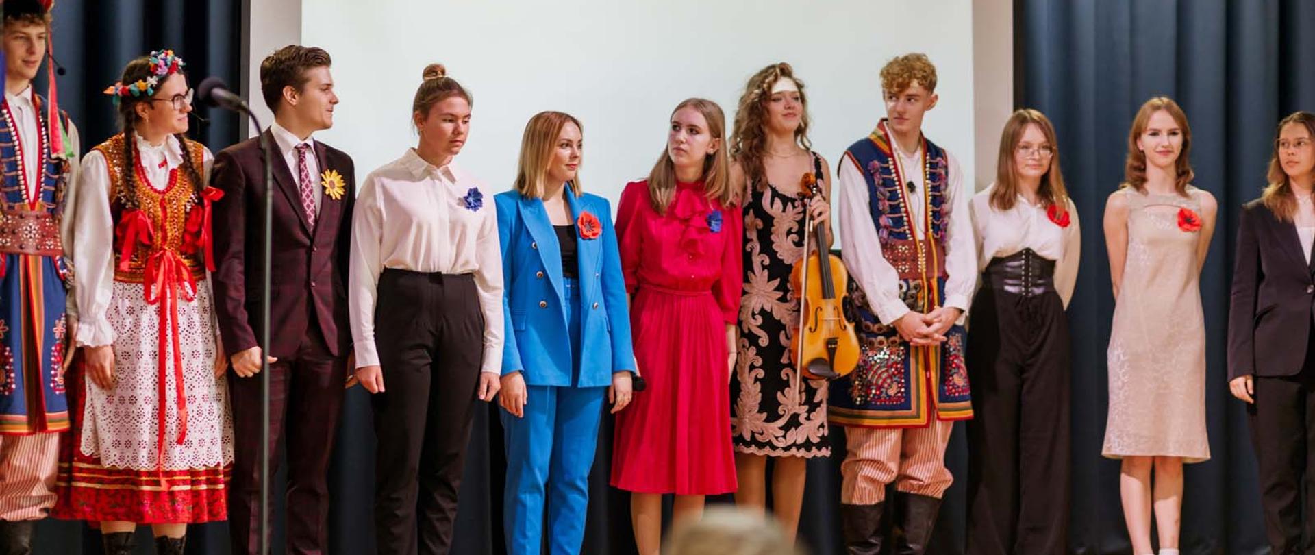 Fotografia przedstawia 11 osób stojących w jednym szeregu na uroczystej Akademii z okazji Jubileuszu szkoły. Część z osób jest ubrana w krakowskie stroje ludowe.