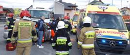 Zdjęcie przedstawia miejsce wypadku. Na pierwszym planie znajdują się strażacy oraz ambulans natomiast na drugim planie widać rozbity samochód marki Mazda 2 oraz samochód ciężarowy. 
