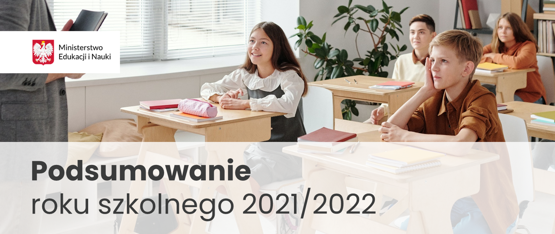 Dzieci słuchające z zaciekawieniem nauczyciela siedzą w klasie w szkolnych ławkach. Poniżej napis: "Podsumowanie roku szkolnego 2021/2022"