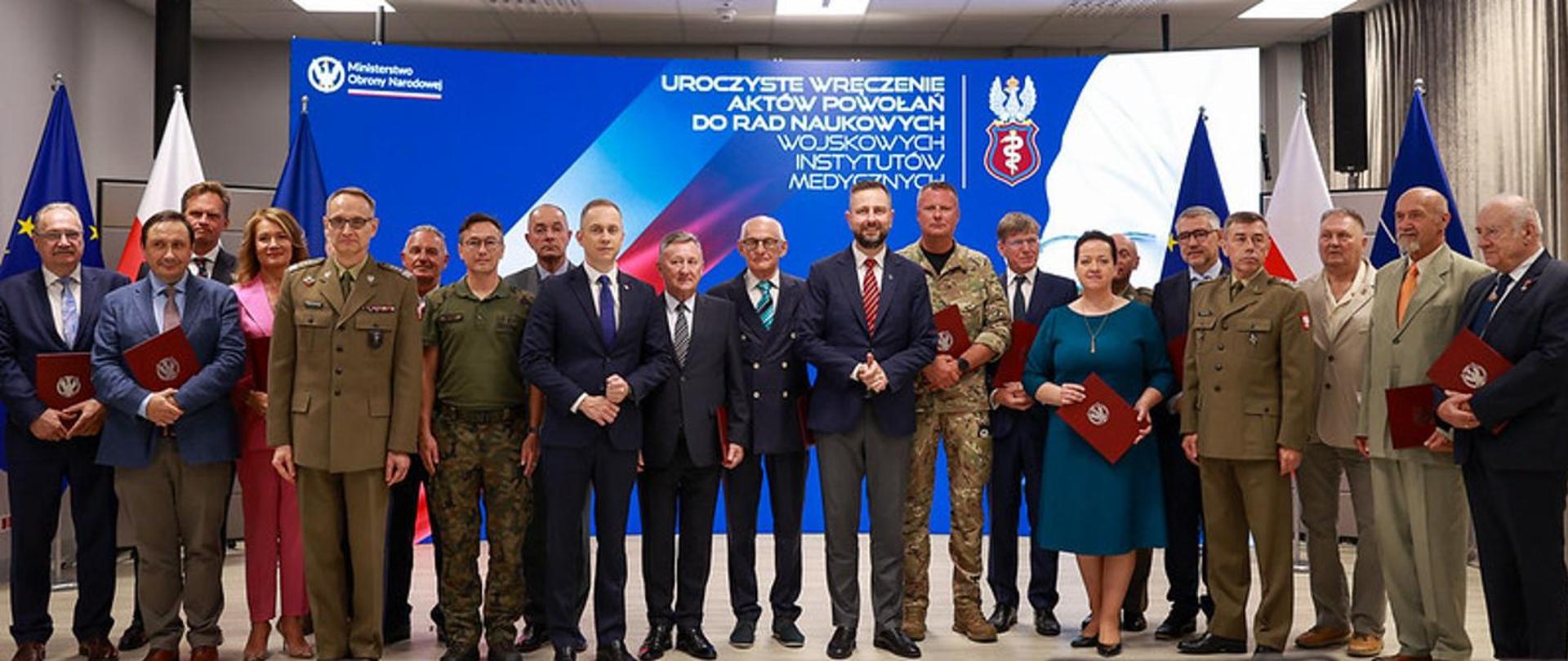 Na zdjęciu widzimy grupę osób podczas uroczystej ceremonii wręczenia aktów powołań do rad naukowych wojskowych instytutów medycznych.
Uczestnicy stoją razem w kilku rzędach, część z nich ubrana jest w mundury wojskowe, a część w garnitury i stroje formalne. Kilka osób trzyma w rękach czerwone teczki z godłem Ministerstwa Obrony Narodowej.
W tle znajduje się duży ekran z napisem:
„Uroczyste wręczenie aktów powołań do rad naukowych wojskowych instytutów medycznych” oraz logotypem Ministerstwa Obrony Narodowej i herbem wojskowej służby zdrowia. Po bokach widać flagi Polski, Unii Europejskiej oraz NATO.
Zdjęcie przedstawia oficjalne wydarzenie państwowe związane z działalnością naukową i medyczną wojska.