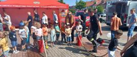 Na zdjęciu widać grupę dzieci przy stanowisku straży pożarnej. Dzieci leją wodę z nalewaków strażackich. Konkurencje nadzorują strażacy w ciemnych strojach. W tle widać czerwony namiot z logo Państwowej Straży Pożarny oraz z białym napisem Komenda Powiatowa Państwowej Straży Pożarnej w Łęczycy. Obok namiotu stoją osoby dorosłe oraz czerwony samochód straży pożarnej. Na zdjęciu widać tył srebrnego samochodu, kamienice oraz drzewa. Jest widno.