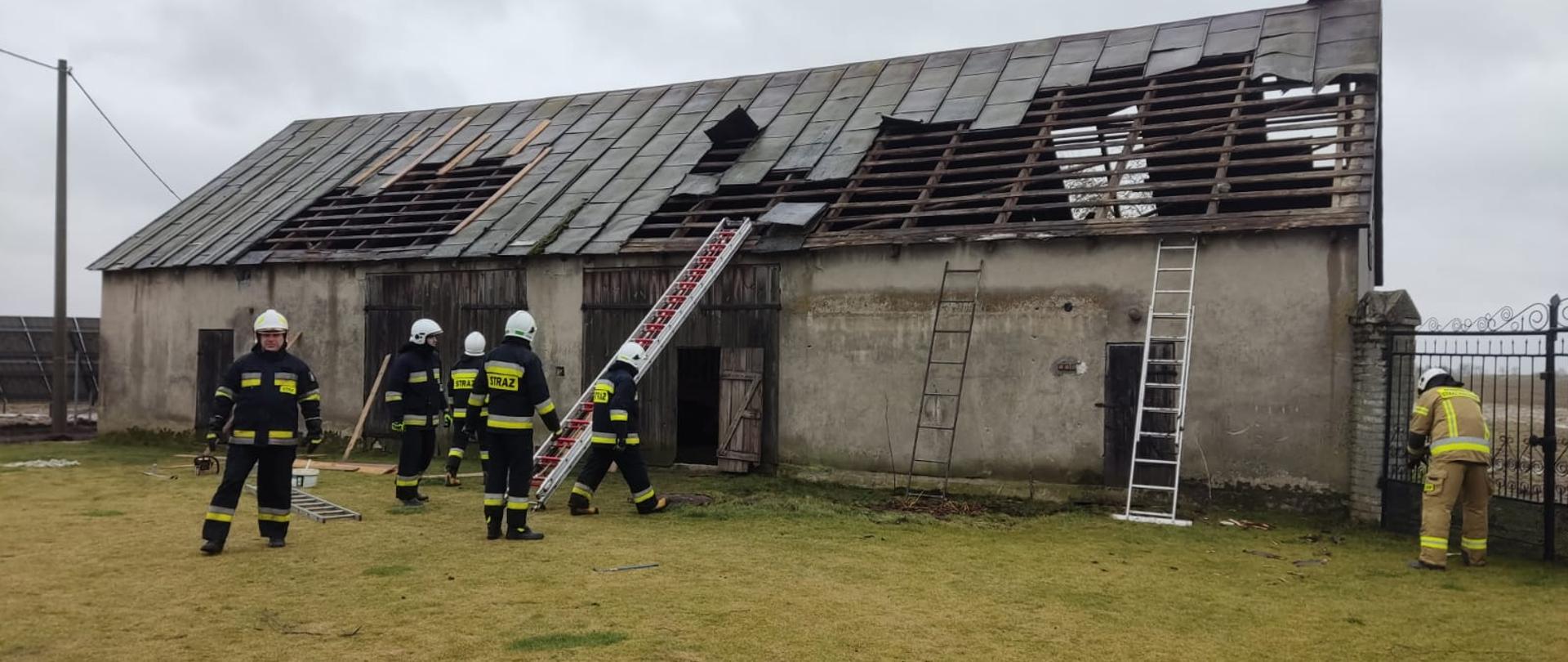Zdjęcie przedstawia budynek gospodarczy z częściowo zerwanym dachem, oraz strażaków z drabinami pożarniczymi w trakcie zabezpieczania dachu przed całkowitym zerwaniem