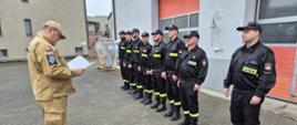 Plac Komendy Powiatowej Państwowej Straży Pożarnej w Sępólnie Krajeńskim, zmiana służbowa ubrana w ubrania koszarowe w szeregu, Komendant Powiatowy Państwowej Straży Pożarnej w Sępólnie Krajeńskim brygadier Marcin Mazurkiewicz w ubraniu służbowym stoi naprzeciw zmiany służbowej i odczytuje rozkaz.