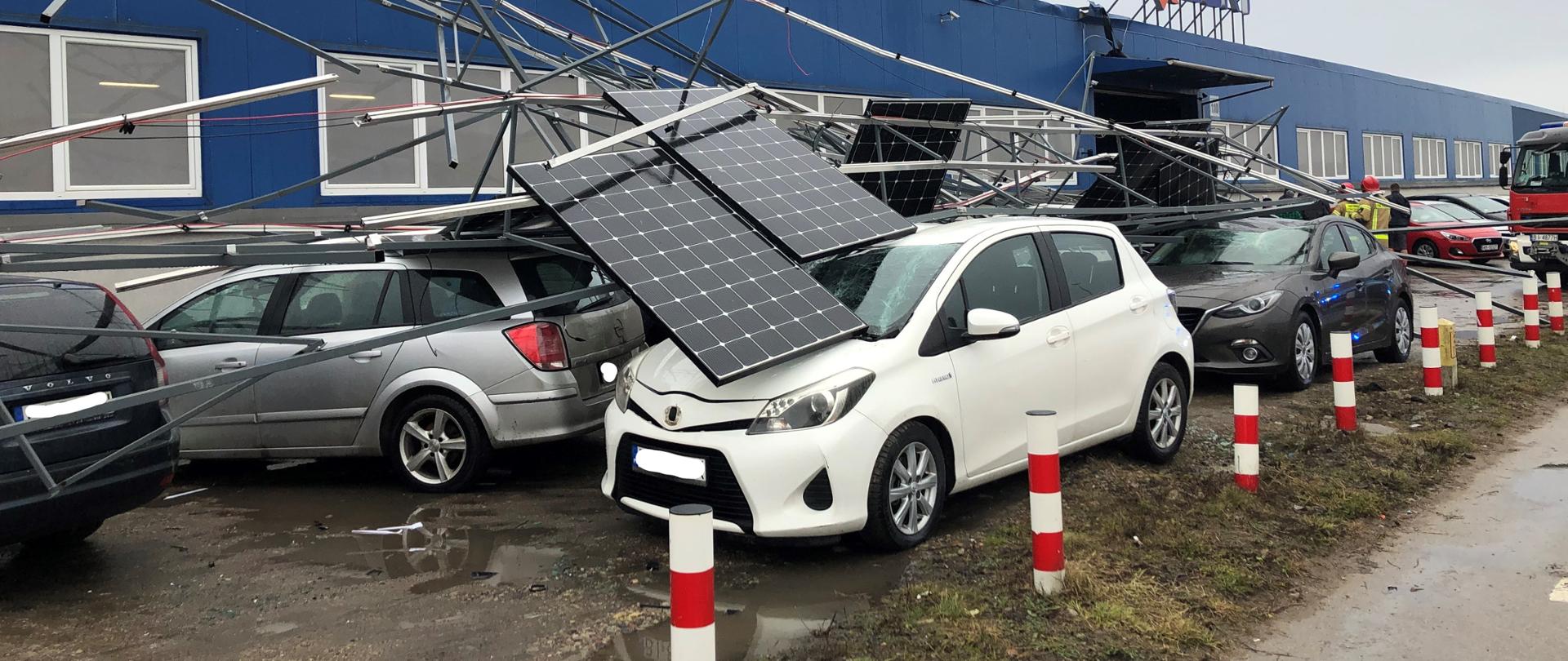Zdjęcie przedstawia pojazdy przygniecione instalacją fotowoltaiczną zerwaną z dachu przez porywisty wiatr