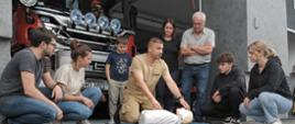 Szkolenie z pierwszej pomocy w remizie strażackiej OSP Rogów. Na pierwszym planie widać grupę siedmiu osób zgromadzonych w kręgu wokół medycznego manekina (fantomu), ułożonego na niebieskiej macie na asfalcie. Strażak w piaskowej koszulce kuca i wykonuje uciski klatki piersiowej na manekinie, instruując pozostałych uczestników, w tym dorośli i młodzież, którzy uważnie obserwują. W tle, w garażu remizy, stoi duży czerwony wóz strażacki z widocznym napisem „OSP ROGÓW” nad przednią szybą i tablicą rejestracyjną SMD 44N1. Budynek ma nowoczesną, betonową fasadę. Ujęcie wykonane jest z niskiej perspektywy.