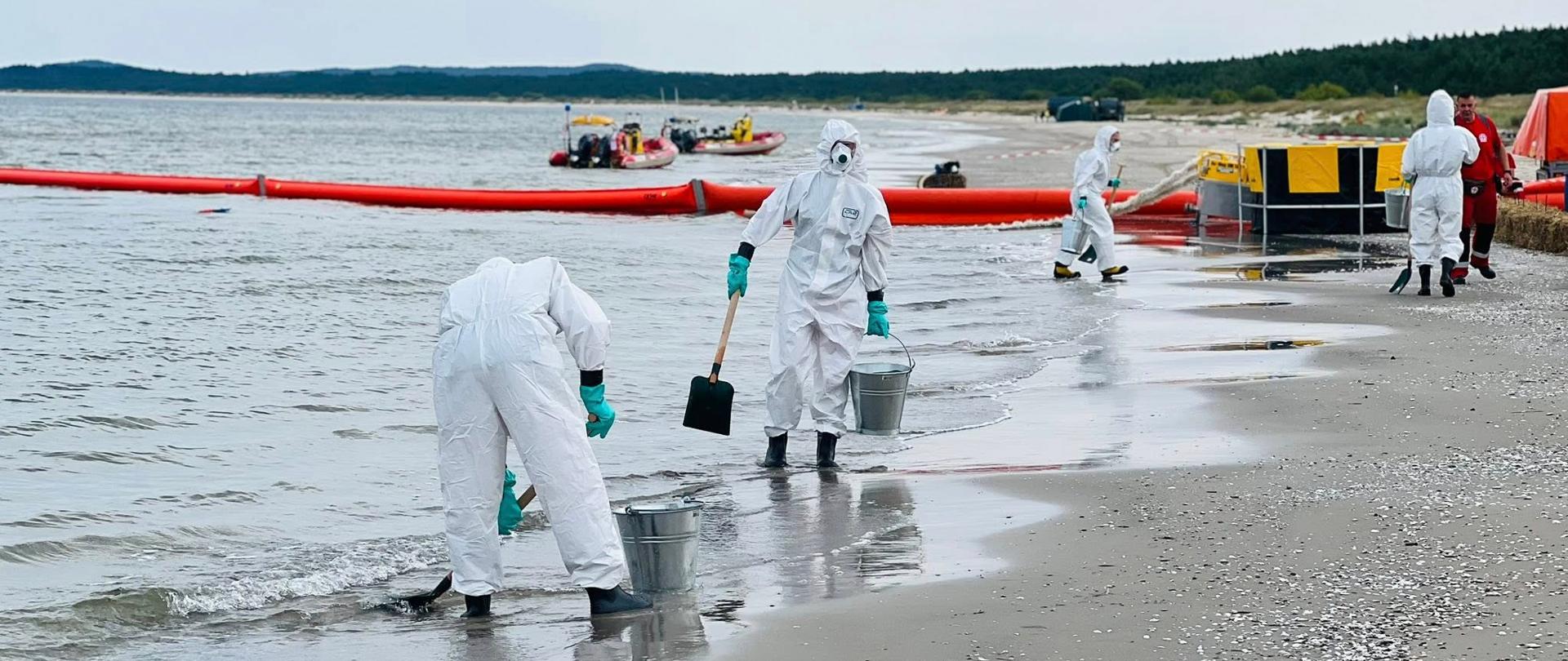 Na zdjęciu widzimy cztery osoby ubrane w białe, ochronne kombinezony. Mają na sobie także maski na twarz i rękawice. Stoją na piaszczystej plaży tuż przy linii brzegowej morza. Woda jest spokojna, a niebo zachmurzone.
Po prawej stronie zdjęcia znajduje się duża, czarno-żółta konstrukcja. Za nią widać dwie osoby, w tym jedną w czerwonej bluzie. Na lewo od tej konstrukcji, na wodzie, unosi się długa, czerwona rura.
Dwóch mężczyzn na pierwszym planie trzyma w rękach wiadra. Jeden z nich, stojący tyłem, pochyla się w stronę wody. Drugi, zwrócony w stronę aparatu, ma w rękach łopatę. W tle, na morzu, widać łodzie motorowe.
