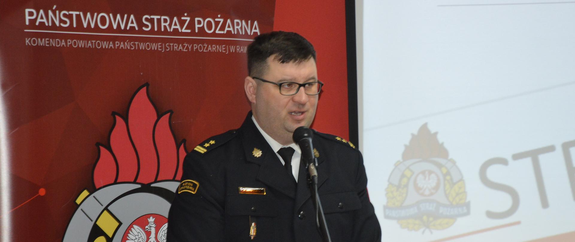 strażak przy mównicy, przemawia, za nm na czerwonym tle logo państwowej strazy pożarnej