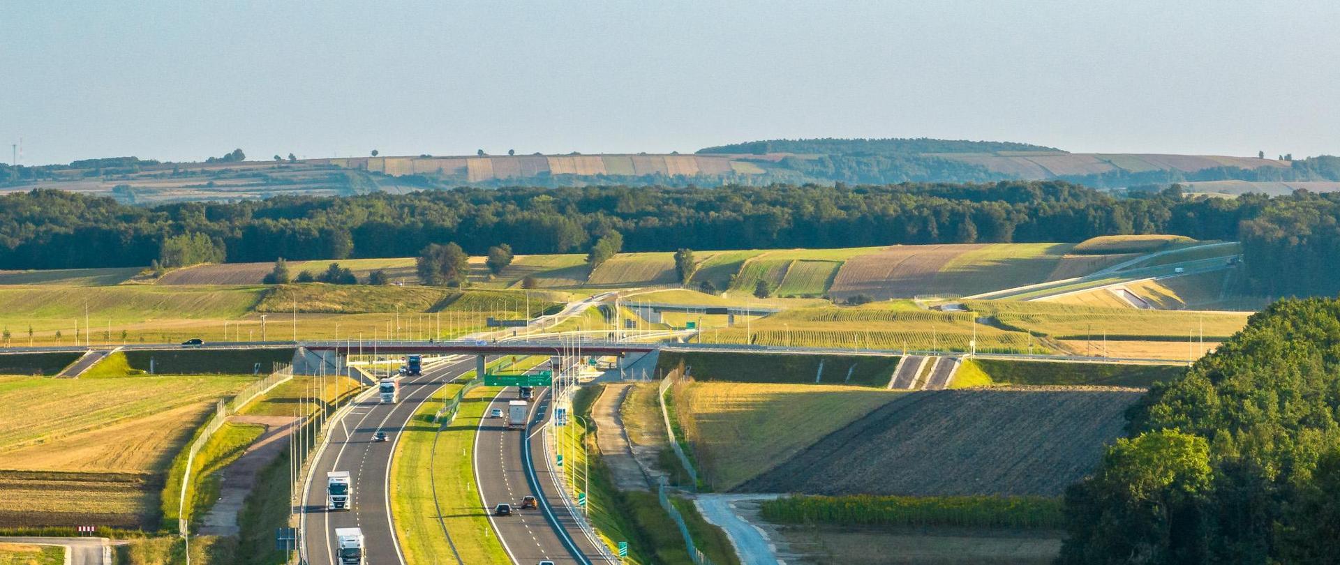Dwujezdniowa droga ekspresowa S7 po dwa pasy ruchu w obu kierunkach. Droga przebiega wśród pól uprawnych i lasów ciągnących się po horyzont.