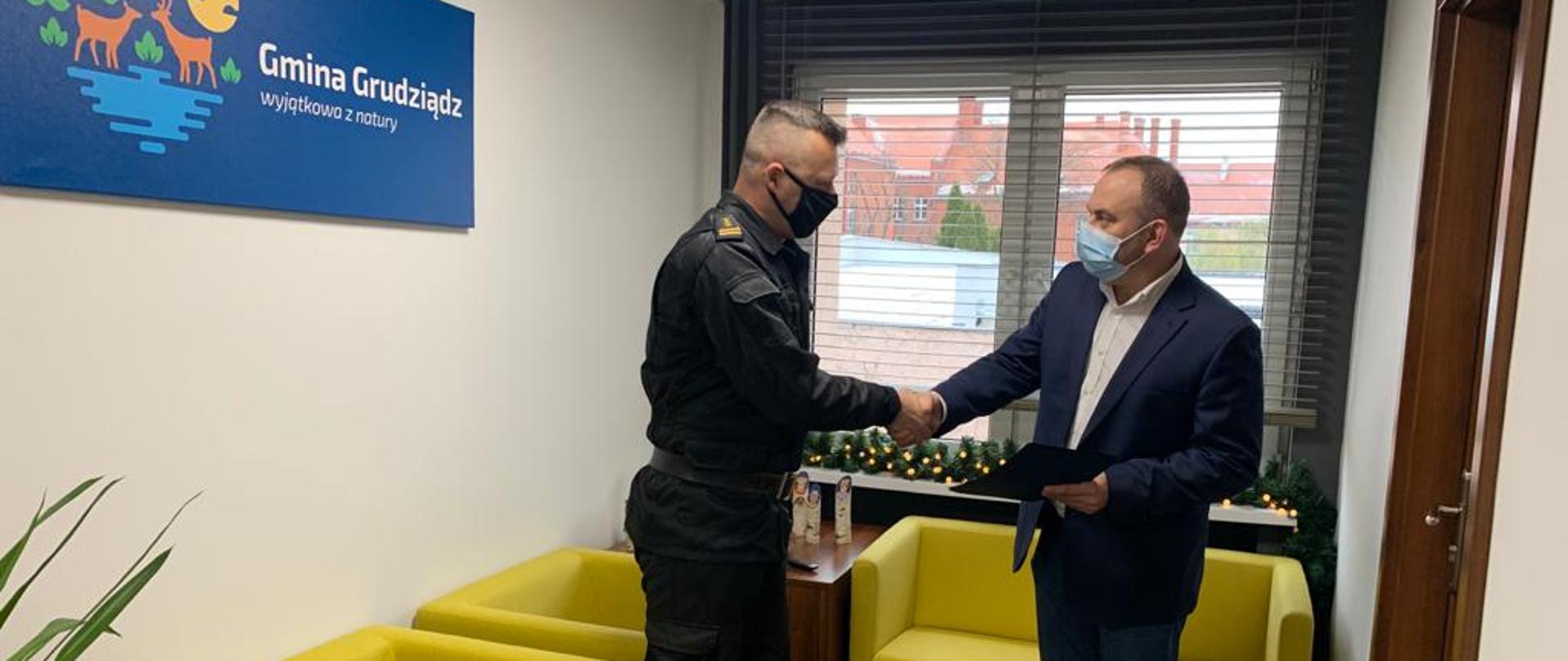Dwaj mężczyźni znajdują się w jasnym w pomieszczeniu. Mężczyzna po lewej stronie w czarnym mundurze, czarnej maseczce i żółto – czarnych butach ściska prawą dłonią dłoń mężczyzny w granatowej marynarce, jasnej koszuli, ciemnych spodniach i niebieskiej maseczce. Mężczyzna po prawej stronie w lewej dłoni trzyma granatową teczkę. W tle trzy fotele w kolorze kanarkowym, plakat z napisem Gmina Grudziądz wiszący na ścianie i okno z żaluzjami i ozdobami świątecznymi na parapecie.