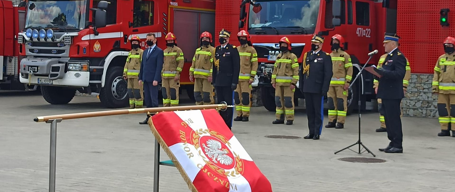 Na pierwszym planie, na stojaku znajduje się sztandar, w tle ustawieni w szeregu strażacy, za nimi pojazdy pożarnicze i bramy garażowe strażnicy.