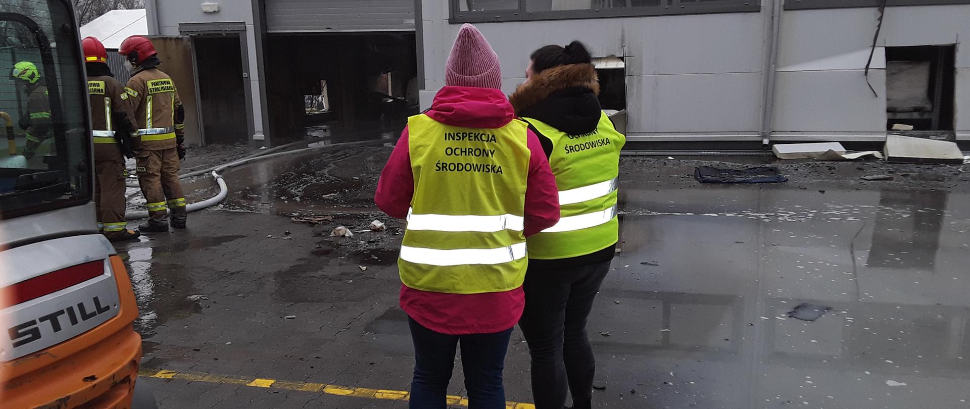 Dwoje inspektorów Wojewódzkiego Inspektoratu Ochrony Środowiska w Warszawie stoi na utwardzonym podłożu i rozpoczyna oględziny miejsca pożaru. W głębi znajduje się hala, która uległa spaleniu.