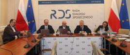 Na zdjęciu widnieje stół prezydialny za którym siedzą od lewej strony: Liwiusz Laska – Dyrektor Generalny w Ministerstwie Rodziny, Pracy i Polityki Społecznej, Sebastian Gajewski - Podsekretarz Stanu w Ministerstwie Rodziny, Pracy i Polityki Społecznej, Norbert Kusiak w zastępstwie za Przewodniczącego zespołu ds. budżetu, wynagrodzeń i świadczeń socjalnych – Leszka Miętka, Jurand Drop – Podsekretarz Stanu w Ministerstwie Finansów oraz Joanna Stachura – Dyrektor w Ministerstwie Finansów i Gospodarki. Po obu stronach stołu widnieją dwie flagi: Polski i UE. W tle siedzących znajduje się baner z napisem Rada Dialogu Społecznego a nad nim wisi godło Polski.