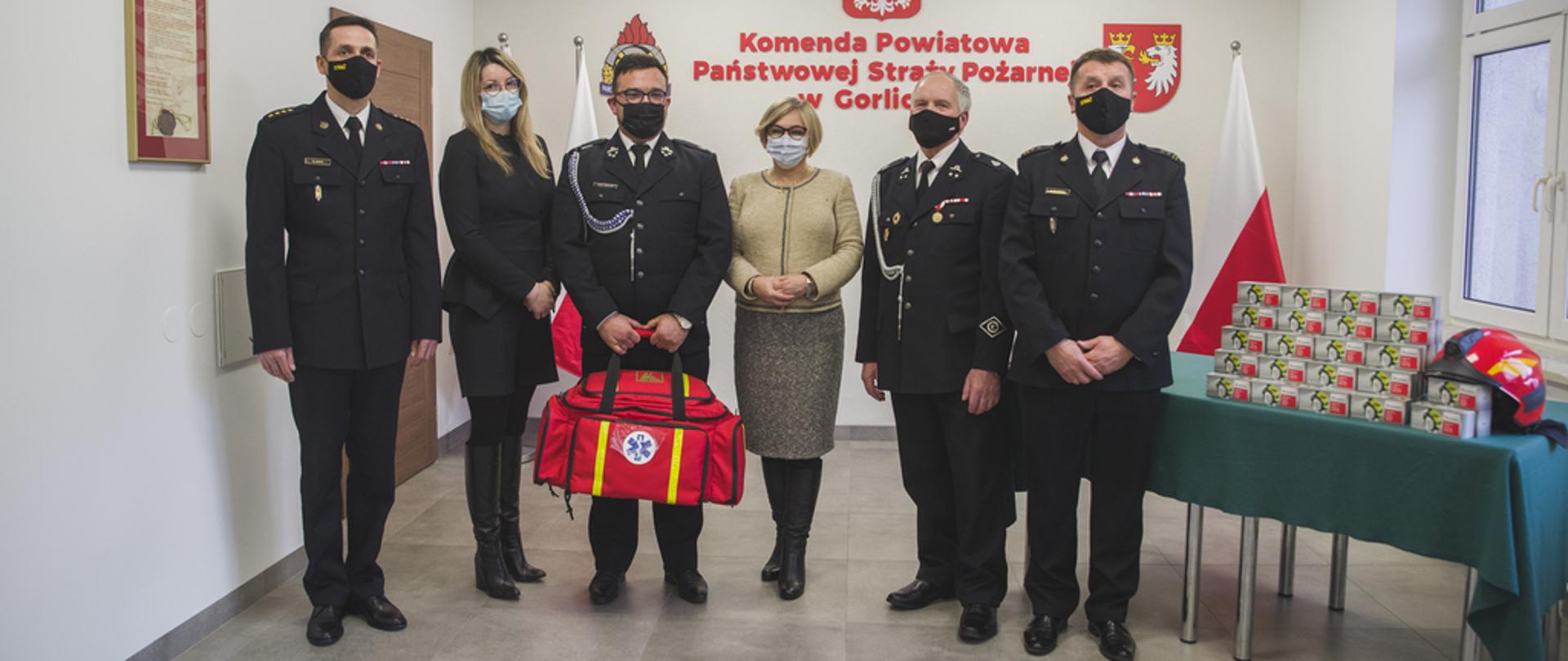 Na zdjęciu strażacy w mundurach wyjściowych, strażacy z Państwowej Straży Pożarnej i z Ochotniczej Straży Pożarnej jeden z nich trzyma torbę R1 do ratownictwa medycznego pomiędzy strażakami stoją w ubraniach cywilnych Pani Poseł i Z-ca Kierownika KRUS.