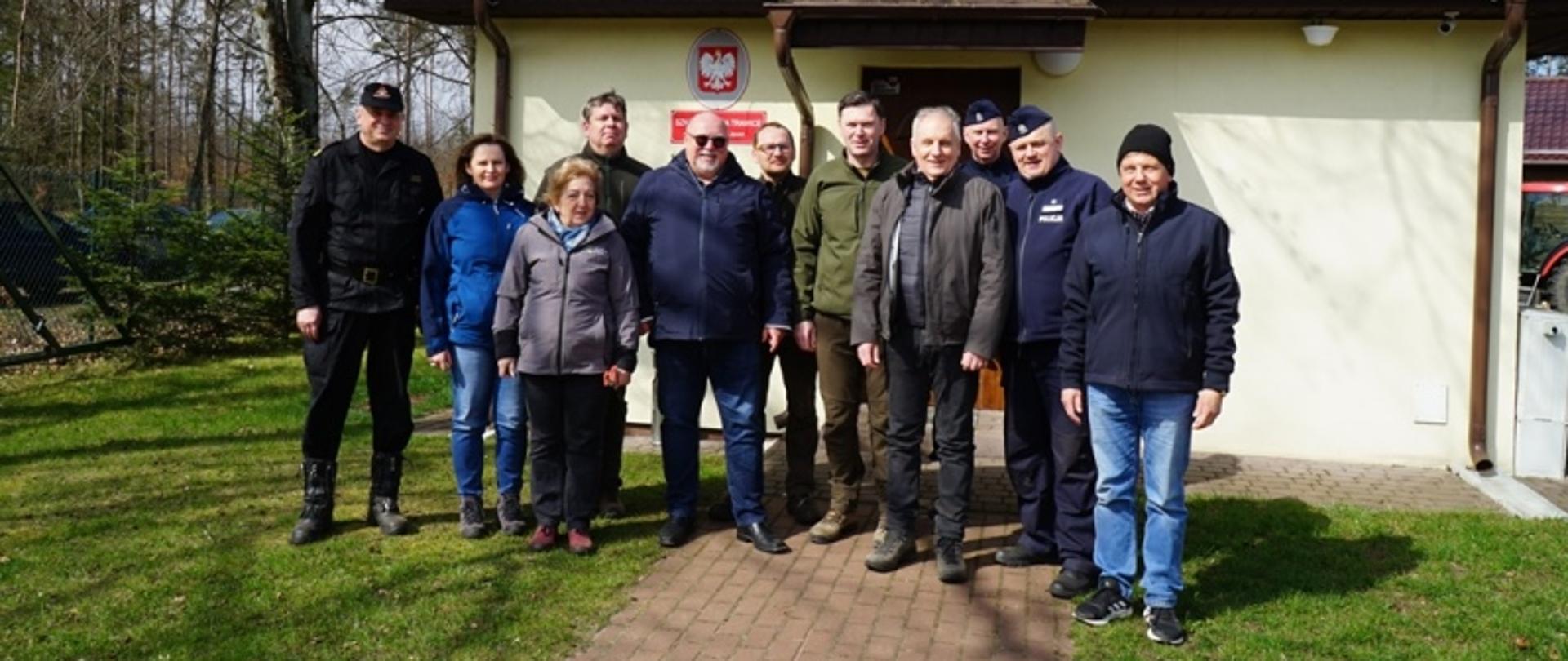 Służby podległe Ministerstwu Spraw Wewnętrznych i Administracji, leśnicy oraz osoby cywilne stoją obok siebie, za nimi znajduje się budynek nadleśnictwa.