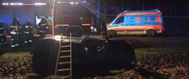 Zdjęcie nocne. Na pierwszym planie leżące na boku auto osobowe. Przy aucie oparta drabina. Auto ma otwarte drzwi zabezpieczone linką przed zamknięciem. Na drugim planie stojące wzdłuż jezdni pojazd gaśniczy i karetka pogotowia. Przy pojeździe gaśniczym grupa strażaków. Całość \oświetlona lampami błyskowymi