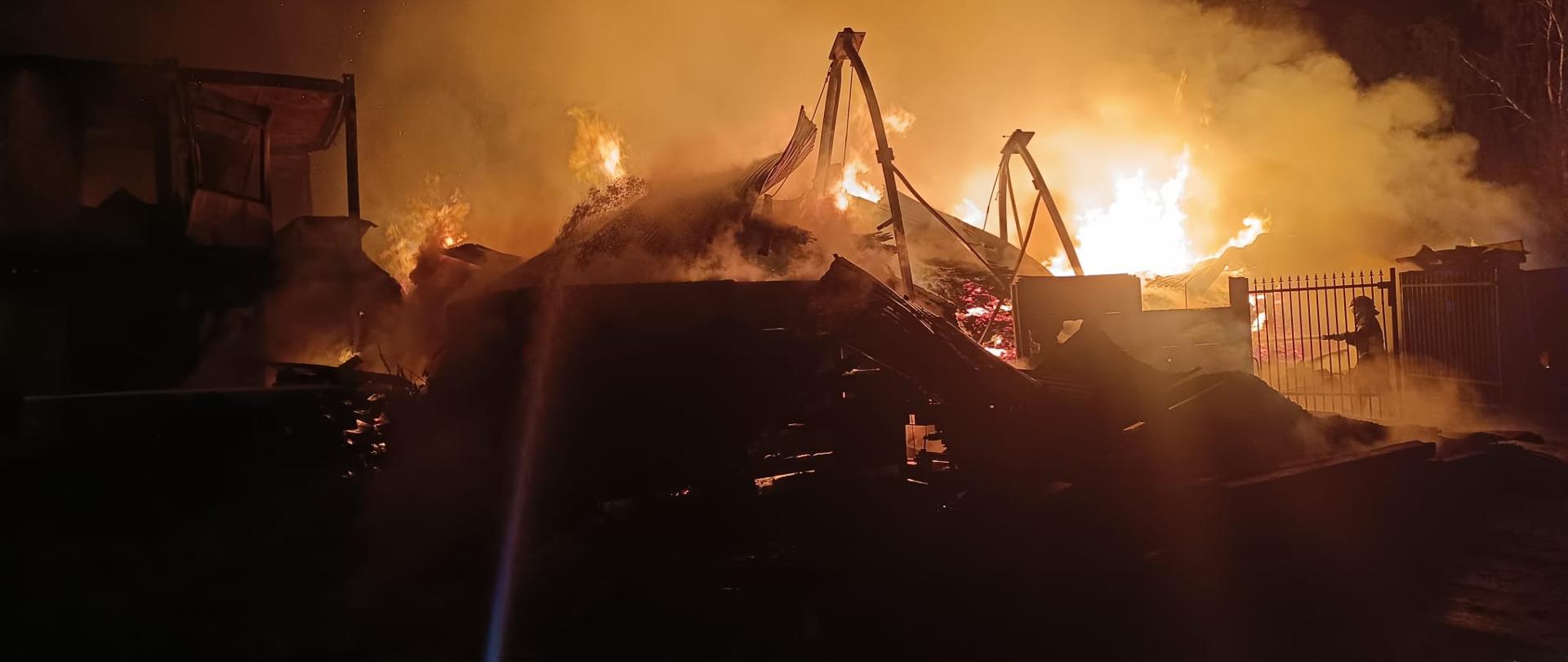 Fotografia przedstawia rozwinięty pożar tartaku. Z prawej strony widać ratownika podającego prąd wody. Na pierwszym planie zniszczona konstrukcja wiaty oraz dym i ogień obejmujący zgromadzony materiał palny.