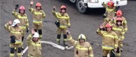 Dziesięciu strażaków stojących wokół odcinka wężowego ułożonego w kształt serca. W tle czerwony samochód pożarniczy