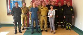 Zdjęcie grupowe wykonane na tle ścianki służbowej. Ścianka czerwona z godłem Polski i logiem Państwowej Straży Pożarnej. W grupie strażacy wraz z rodzicami Piotra Milczarka.