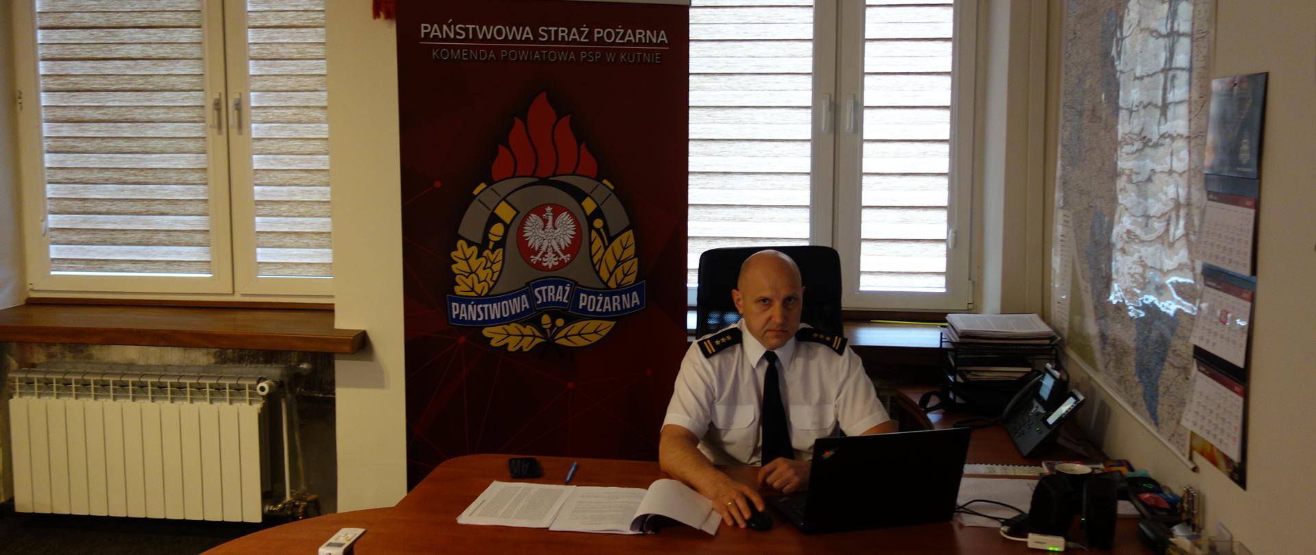 Pomieszczenie gabinetu komendanta Powiatowego PSP w Kutnie. Osoba Komendanta w białej koszuli wyjściowej prowadzi przy użyciu laptopa wideokonferencję. W tle za osobą komendanta baner z logo Państwowej Straży Pożarnej.