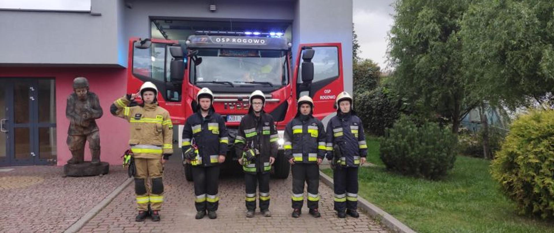 Zdjęcie wykonane przed remizą OSP Rogowo. Przed budynek stoją strażacy.Część ma mundur piaskowy, część ciemny. Strażacy stoją w szeregu. Mają białe hełmy.