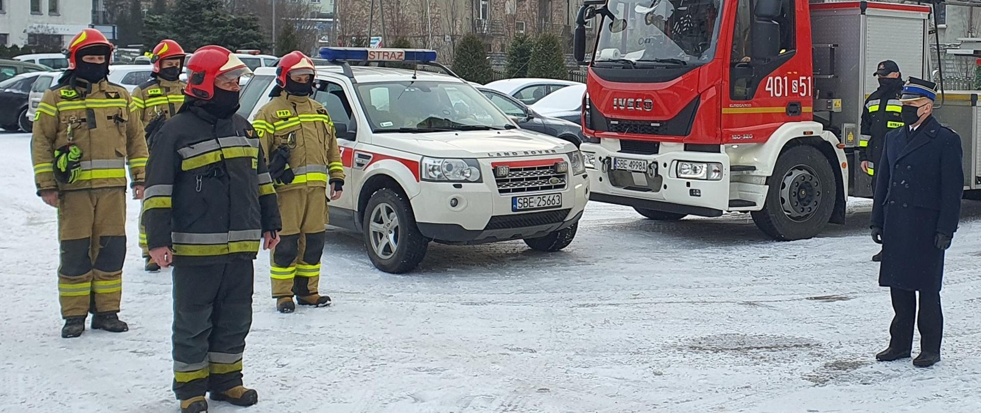 Zdjęcie wykonane na placu Komendy Powiatowej Państwowej Straży Pozarnej na tle dówch samochodów pożarniczych. Komendant Powiatowy mł. bryg. Arkadiusz Spera oraz strażacy Jednostki Ratowniczo - Gaśniczej ubrani w ubranie specjalne żegnają odchodżcego na emeryturę st. ogn,. Tomasza Gajka. 