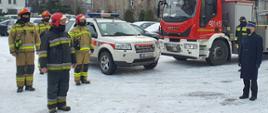 Zdjęcie wykonane na placu Komendy Powiatowej Państwowej Straży Pozarnej na tle dówch samochodów pożarniczych. Komendant Powiatowy mł. bryg. Arkadiusz Spera oraz strażacy Jednostki Ratowniczo - Gaśniczej ubrani w ubranie specjalne żegnają odchodżcego na emeryturę st. ogn,. Tomasza Gajka. 