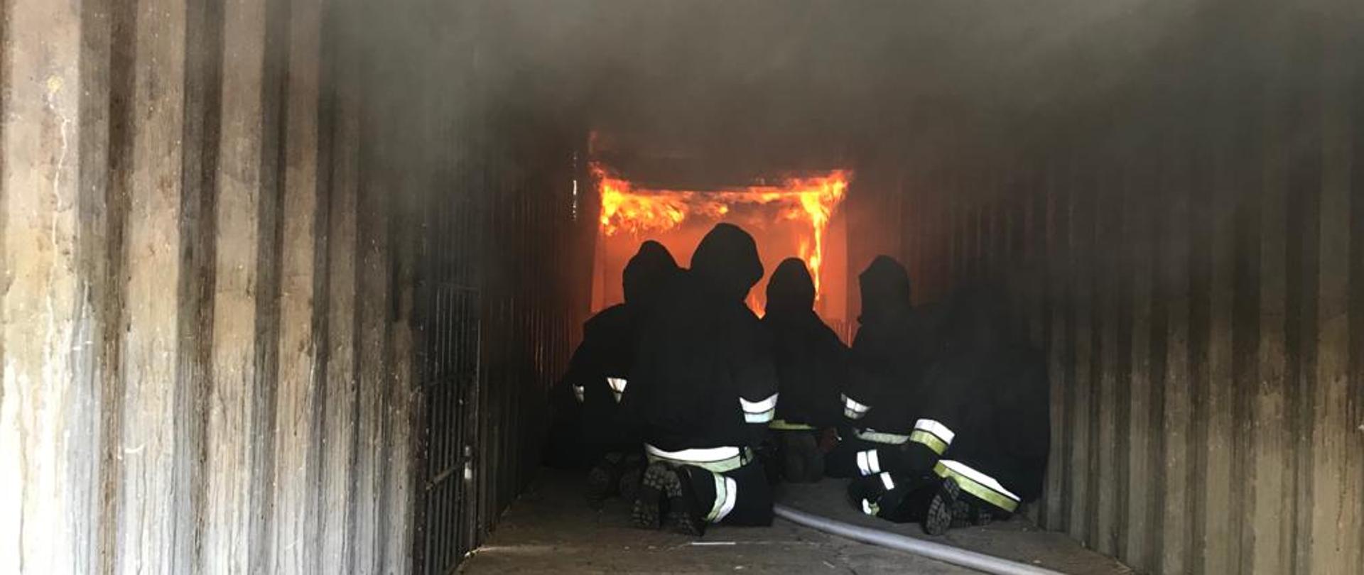 Strażacy przebywający w środku komory ogniowej. W tle widoczne płomienie.