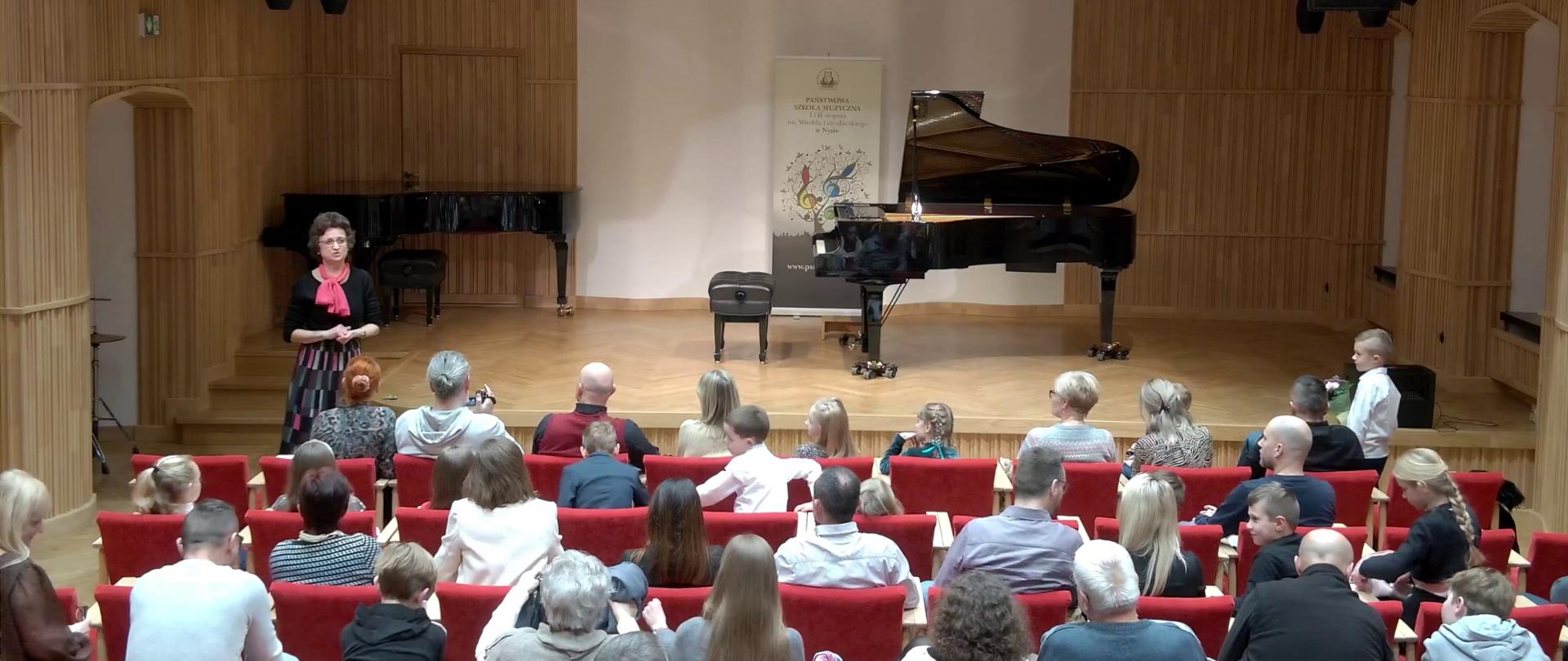 Zdjęcie przedstawia salę koncertową PSM w Nysie podczas koncertu uczniów klas fortepianu. W dolnej części słuchacze na widowni, zapowiada pani Bogusława Kos. W głębi estrada, na której stoi otwarty czarny fortepian. Zdjęcie jest częścią albumu zdjęć z koncertu, który odbył się 13 grudnia 2022..