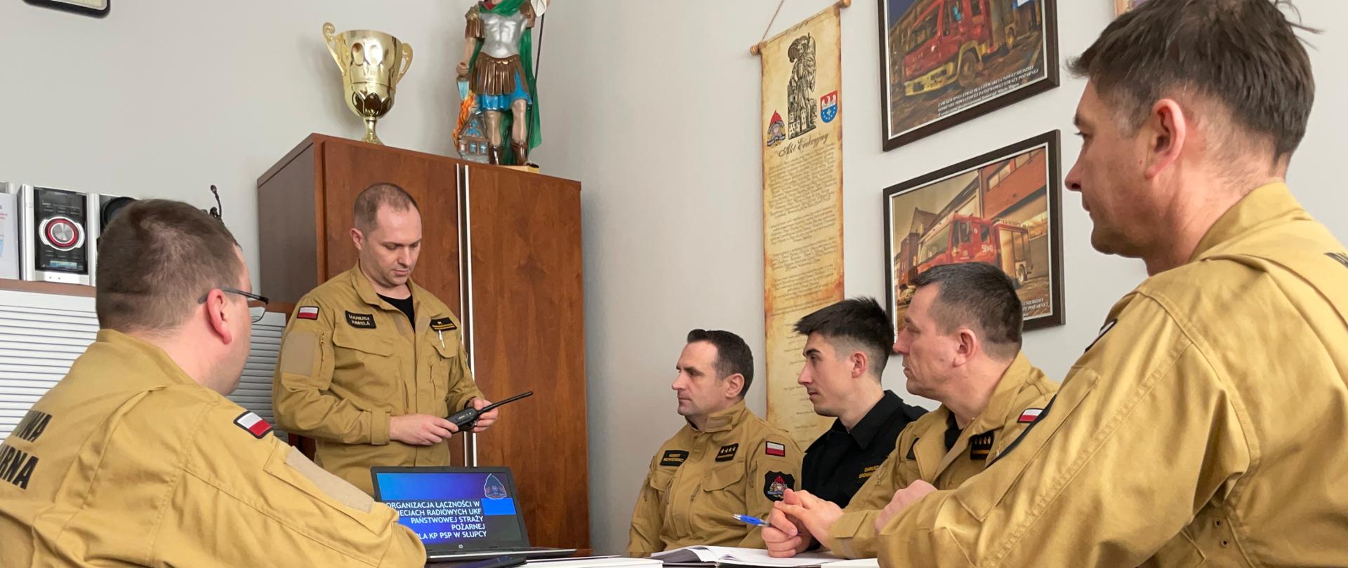 sześciu funkcjonariusz siedzi w pomieszczeniu podczas szkolenia na stole laptop przed nim stoi jeden funkcjonariusz i trzyma w ręku radiotelefon 