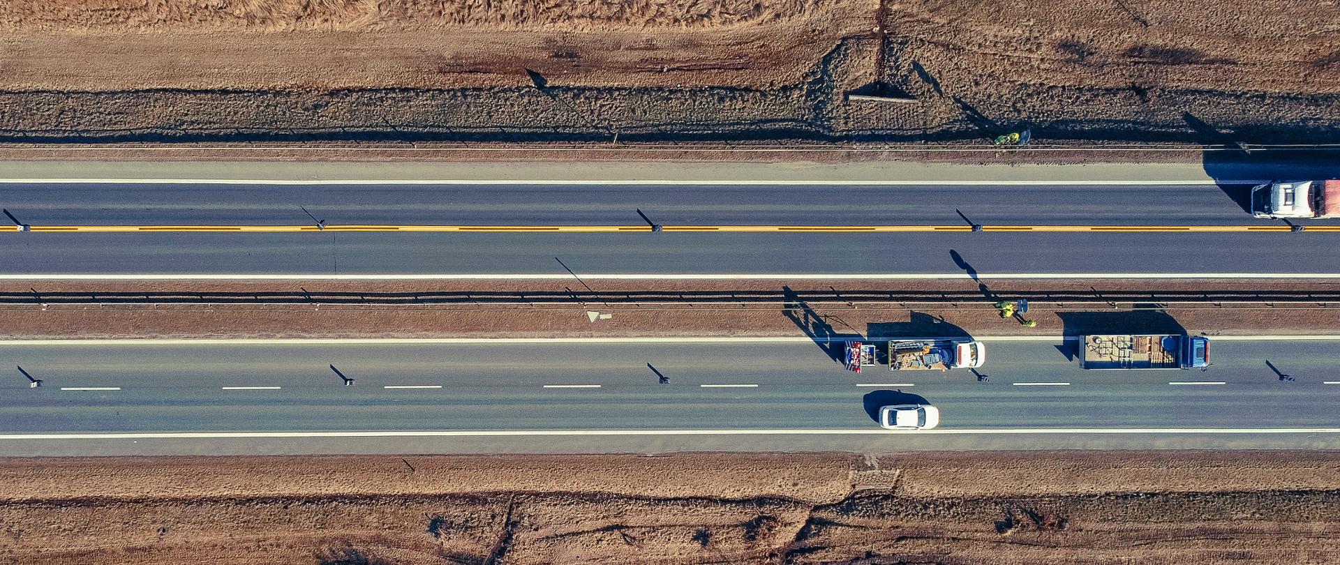 Zdjęcie z lotu ptaka dwujezdniowej drogi przebiegającej przez fotografie horyzontalnie. Na fotografii widoczna brygada pracowników wprowadzającą nową organizację ruchu. Na wyżej położonej jezdni widoczna podwójna ciągła lina w kolorze żółtym. 