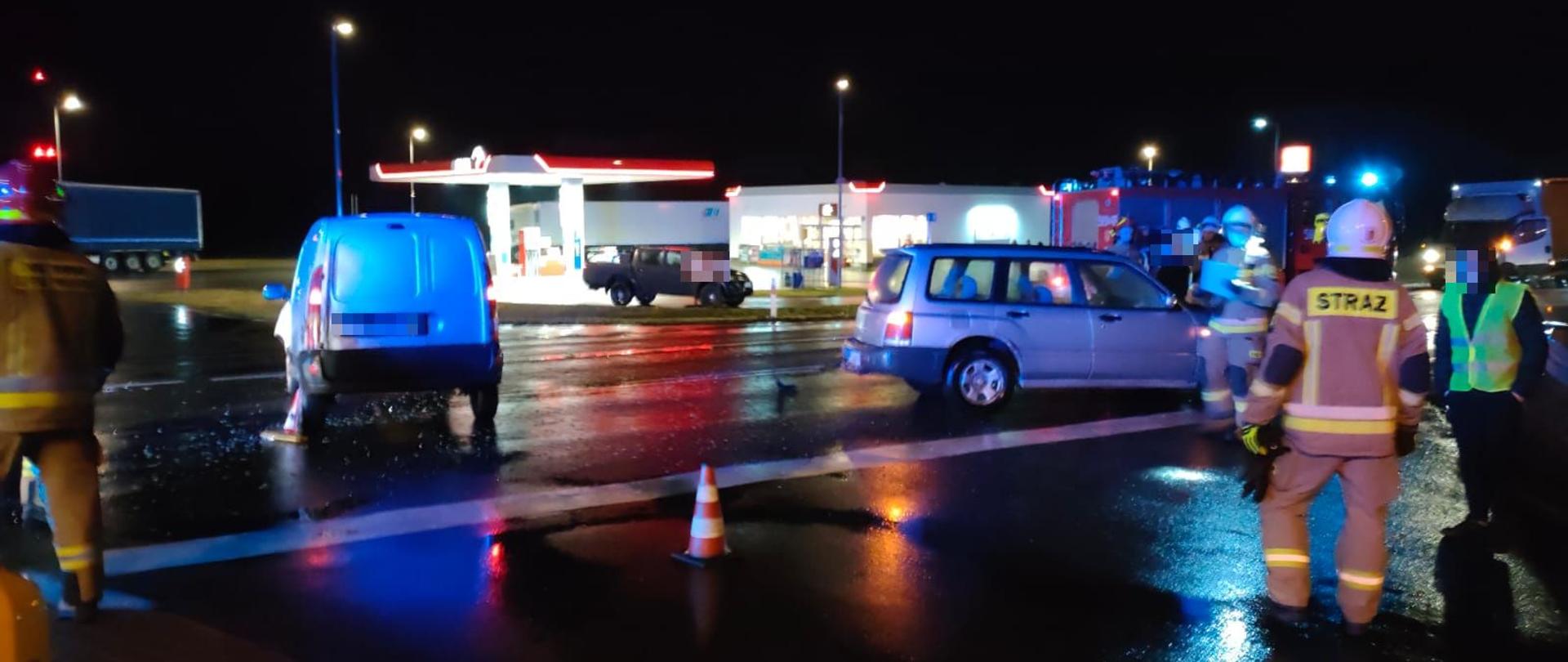 Dwa srebrne samochody rozbite na środku drogi w tle widać stacje benzynową. Na pierwszym planie z prawej i lewej strony stoją strażacy oraz inna osoba w żółtej kamizelce