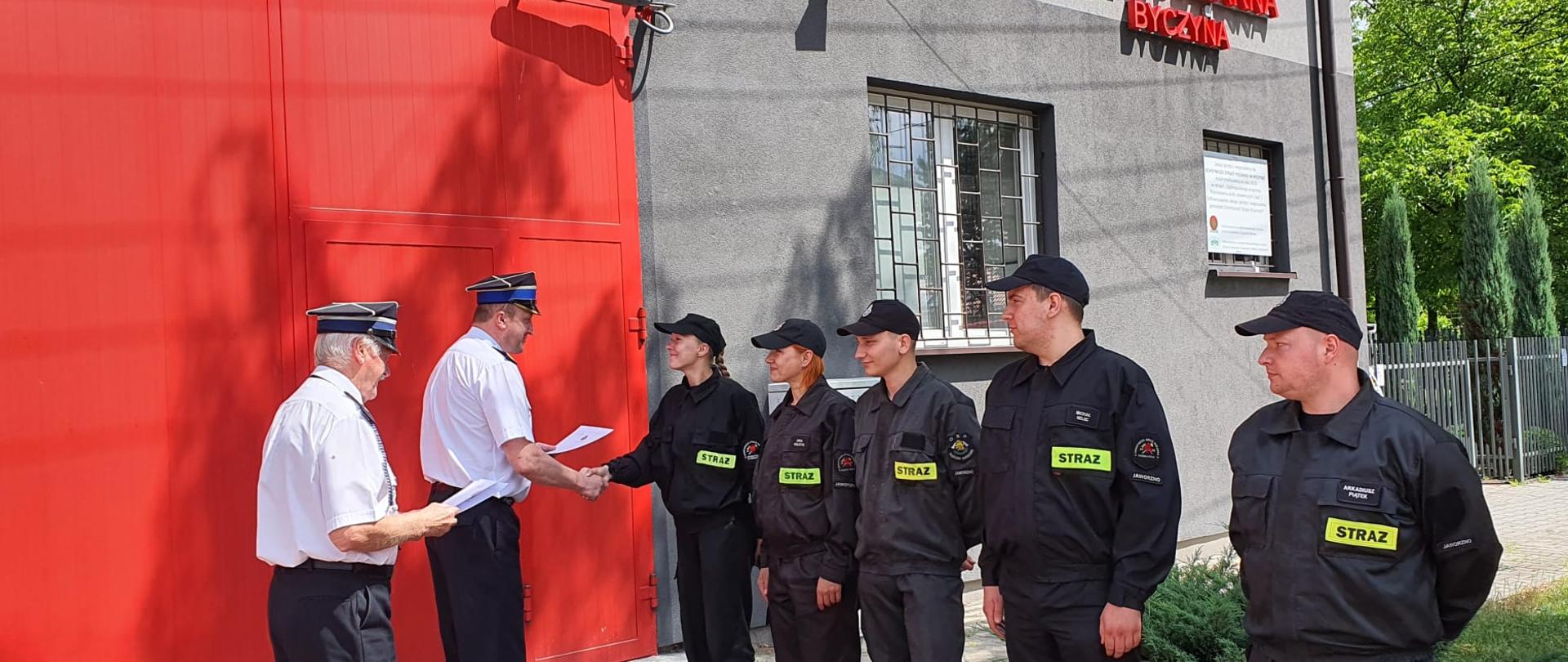 Zdjęcie przedstawia komendanta miejskiego PSP w Jaworznie wręczającego świadectwa ukończenia szkolenia kierujących działaniem ratowniczym dla członków OSP z terenu powiatu na tle jednostki OSP Byczyna.