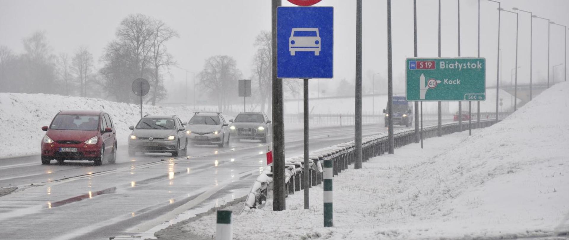 Zima. Widok z poziomu jezdni. Samochody jadące drogą. Na pierwszym planie znak informujący o wjeździe na drogę ekspresową.