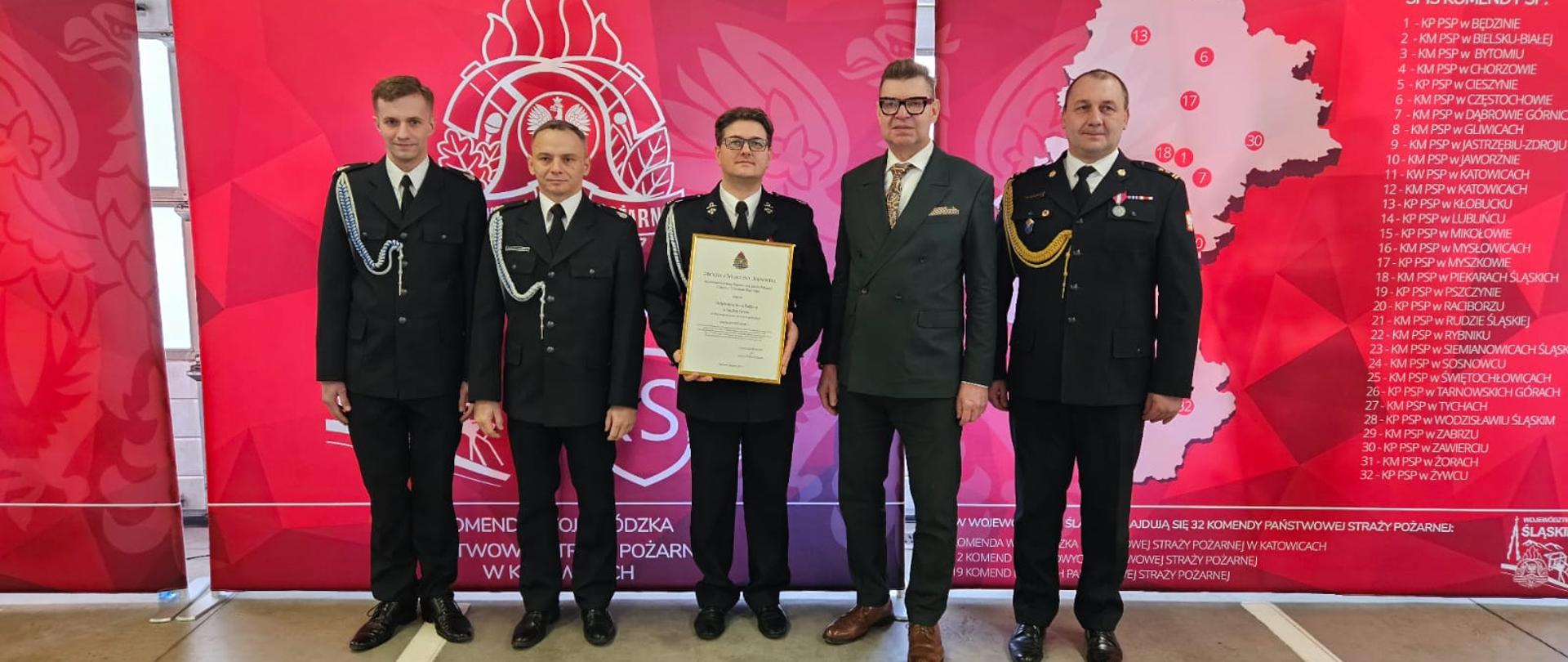 Na pierwszym planie przedstawiciele Ochotniczej Straży Pożarnej w Suchej Górze w obecności Sekretarza Miasta Bytom Pana Mirosława Luksa i Komendanta Miejskiego PSP w Bytomiu bryg. Tomasza Rutkowskiego. Czerwone banery Państwowej Straży Pożarnej w tle. 